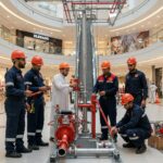 ### Key Elements**Main Subjects**  A group of six individuals, including five in blue work uniforms and helmets, and one in traditional white attire.**Objects**  A fire safety demonstration setup featuring hoses, pumps, and a tall vertical structure.**Notable Features**  The individuals are engaged in a collaborative task, with tools and equipment indicative of fire safety training.**Text/Symbols**  Presence of safety helmets and work uniforms suggests a professional and safety-oriented environment.### Overall Mood/Theme  The image conveys a sense of teamwork and professionalism in a safety training context, highlighting the importance of fire safety awareness in public spaces.