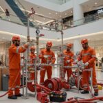 ### Key Elements

**Main Subjects**  
- Five workers in orange safety suits and helmets.

**Objects**  
- A complex assembly of pipes and valves.
- Firefighting equipment, including pumps and hoses.
- Tools and equipment on a workbench.

**Notable Features**  
- Indoor setting, likely a mall or commercial space.
- Modern architecture in the background.
- Safety gear prominently displayed.

**Text/Symbols**  
- Company branding on uniforms and equipment.

### Overall Mood/Theme  
The image conveys a theme of teamwork and professionalism in a safety-focused environment. The workers appear engaged and focused on their task, suggesting a sense of urgency and importance related to fire safety and preparedness. The bright colors of their uniforms contrast with the sleek, modern backdrop, emphasizing the industrial aspect within a commercial space.