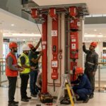 ### Key Elements

- **Main Subjects**: Six individuals in safety gear (hard hats and reflective vests) working on a fire safety system.
- **Objects**: Fire safety equipment, including pipes, alarms, and a ladder.
- **Notable Features**: Red pipes and alarms prominently displayed; safety equipment emphasizes the importance of fire safety.
- **Text/Symbols**: Safety labels and signs indicating fire safety procedures.

### Overall Mood/Theme
The scene conveys a sense of urgency and professionalism, highlighting teamwork and safety in a commercial environment. The focus on fire safety equipment underscores the importance of preparedness and compliance in public spaces.